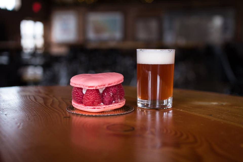 A Raspberry Macaroon made by Black Cat Bake Shop perched to the left of a small 4oz pour of a Draught Works sour beer. 